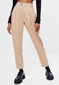 Beige skräddarsydda byxor med veck, hög midja och knappstängning. Bärs med kraftiga svarta skor och ribbad svart långärmad topp.