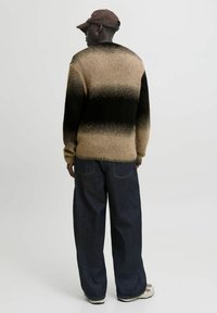 Mann mit einer braunen Mütze, einem braun-schwarz verlaufenden Strickpullover, weiten dunklen Jeans und weißen Sneakers, der mit dem Rücken zur Kamera steht.