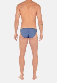 Mann steht barfuß, Rückenansicht, trägt einfarbige blaue Badehose, mit einem einzigen Band-Tattoo am linken Unterarm vor schlichtem weißem Hintergrund.