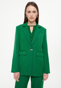 Blazer verde su misura con rever affilati, due tasche frontali e una chiusura in tono oro. Indossato su un top verde con scollo a chiave.