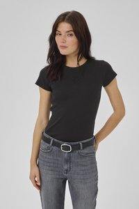 Tumepruuni juustega naine, kes kannab kitsast musta lühikeste varrukatega pluusi ja kõrge vöökohaga halli teksaseid musta vööga, seistes halli tausta ees.