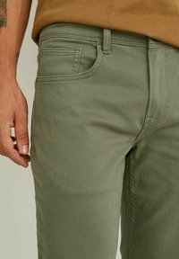 Ljus olivgröna byxor i bomull med en något texturerad yta, framfickor och en avslappnad passform, visade på en models sida.