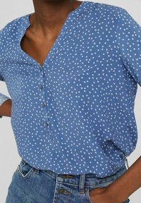 Una camicia azzurra con bottoni, caratterizzata da un collo a V, pois bianchi e maniche lunghe, realizzata in un tessuto leggero. Etichetta di lavaggio visibile.