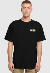 Sort bomulds T-shirt med korte ærmer, der præger "HAWAI" og "E KOMO MAI" i lys beige tekst øverst til venstre.