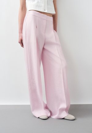 Donna che indossa pantaloni larghi rosa chiaro e un top bianco corto senza maniche, in piedi contro un muro bianco semplice.