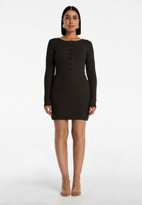 Sukienka z długim rękawem, w ciemnym brązowym kolorze, z okrągłym dekoltem, zapinana na guziki z przodu, o dopasowanej sylwetce, stylizowana z przezroczystymi sandałami na obcasie.