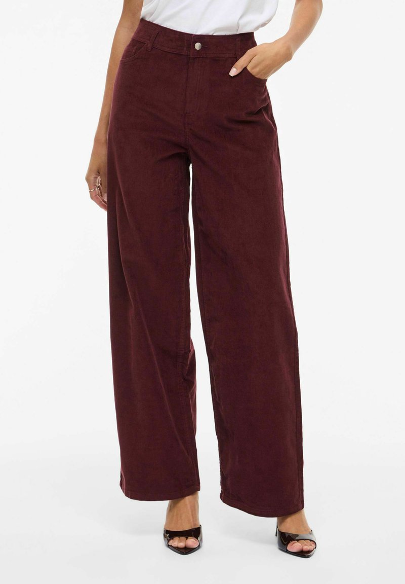 Wijd uitlopende bordeauxrode ribfluwelen broek met hoge taille, riemlussen, zijzakken en een knoopsluiting aan de voorkant; gladde textuur met een lichte glans.