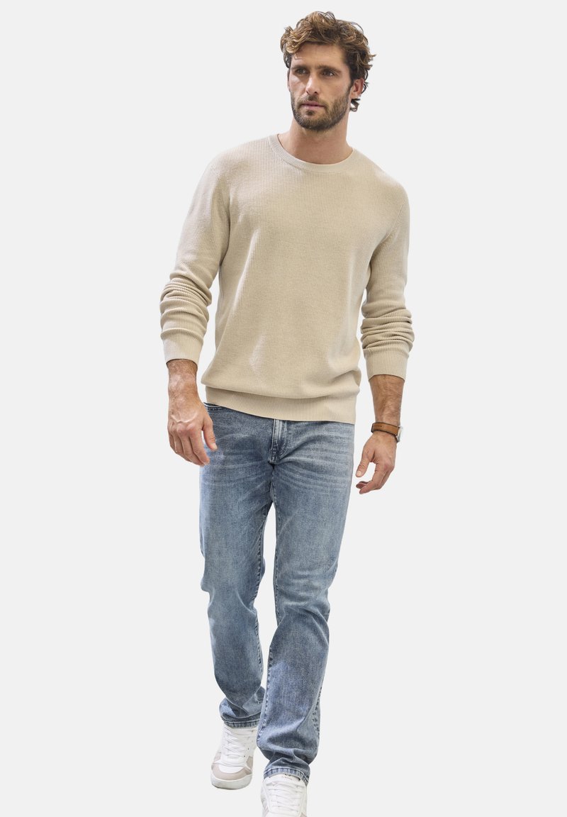 Beige strukturierter Strickpullover mit Rundhalsausschnitt, kombiniert mit hellblauen Jeans und weißen Sneakers. Model, das vor einem schlichten Hintergrund läuft.