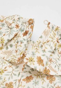 Acercamiento de una tela crema con estampado floral en verde, naranja y amarillo, mostrando botones de madera y lazos de botones en secciones fruncidas.