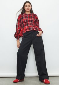 Top peplum a quadri rossi con maniche lunghe a palloncino, abbinato a pantaloni neri a gamba larga e vita alta e scarpe rosse con dettagli bianchi.