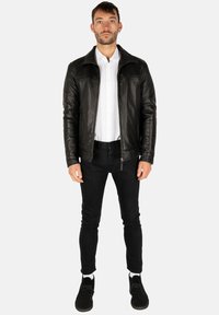 Giubbotto di pelle nera con colletto a rever, chiusura con zip e due tasche laterali, indossato sopra una camicia bianca e jeans skinny neri con scarpe nere.