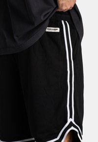 Shorts de sport noirs avec des rayures blanches sur les côtés et un tissu en maille texturée. Une étiquette "PEGADOR" est visible près de la taille.