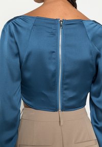 Top cropped em cetim azul com um decote largo e mangas longas; apresenta um fecho de correr prateado visível nas costas e uma textura suave.