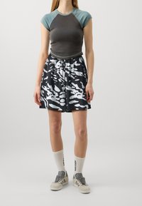 Femme portant un t-shirt ajusté bicolore, une jupe à motifs noirs et blancs, des chaussettes blanches montant jusqu'à la cheville avec du texte, et des baskets blanches et grises, debout.