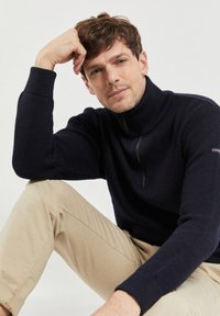 Granatowy sweter zapinany na zamek o rypsowym wzorze, z wysokim kołnierzem i kontrastowym logo na rękawie, zestawiony z jasnobeżowymi spodniami.