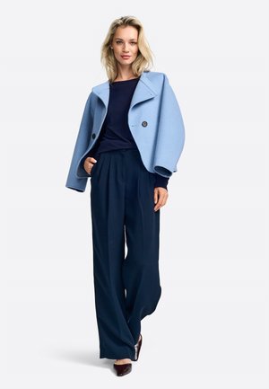Cappotto di lana azzurro chiaro con un ampio colletto e due bottoni neri, abbinato a pantaloni ampi blu scuro e scarpe scure.
