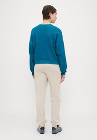 Felpa teal con maniche ampie, abbinata a pantaloni felpati beige. Entrambi i capi hanno una vestibilità comoda e una texture morbida. Sneakers grigie visibili.