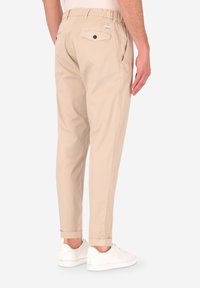 Chinos in cotone beige con vita elastica, due tasche posteriori con chiusura a bottone e orli arrotolati. Indossati con sneakers bianche.