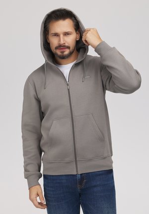 Mężczyzna z brodą w szarej bluzie na suwak i niebieskich dżinsach, przeciąga kaptur na głowę jedną ręką.