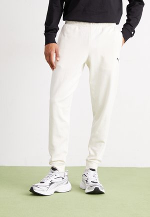 Joggers blancos de algodón con cintura elástica, combinados con zapatillas deportivas en blanco y negro. El fondo presenta un suelo verde.