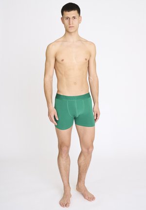 Jonge man die blootsvoets staat en groene boxerbriefs draagt tegen een effen lichte achtergrond, voor zich uitkijkend.