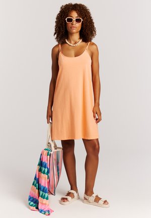 Vrouw met krullend haar, gekleed in een perzikkleurige zomerse jurk, beige sandalen, zonnebril, kralenhalsketting, en met een gestreepte boodschappentas met kleurrijke handdoek.