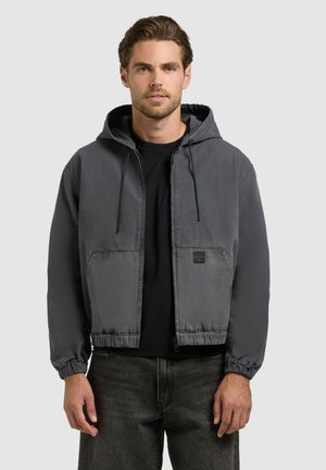 Muž nosiaci kapucňovú bundu v antracitovej sivej farbe s prednými vreckami, pod ktorou má čierne tričko a tmavé džínsy, stojaci pred svetlosivým pozadím.