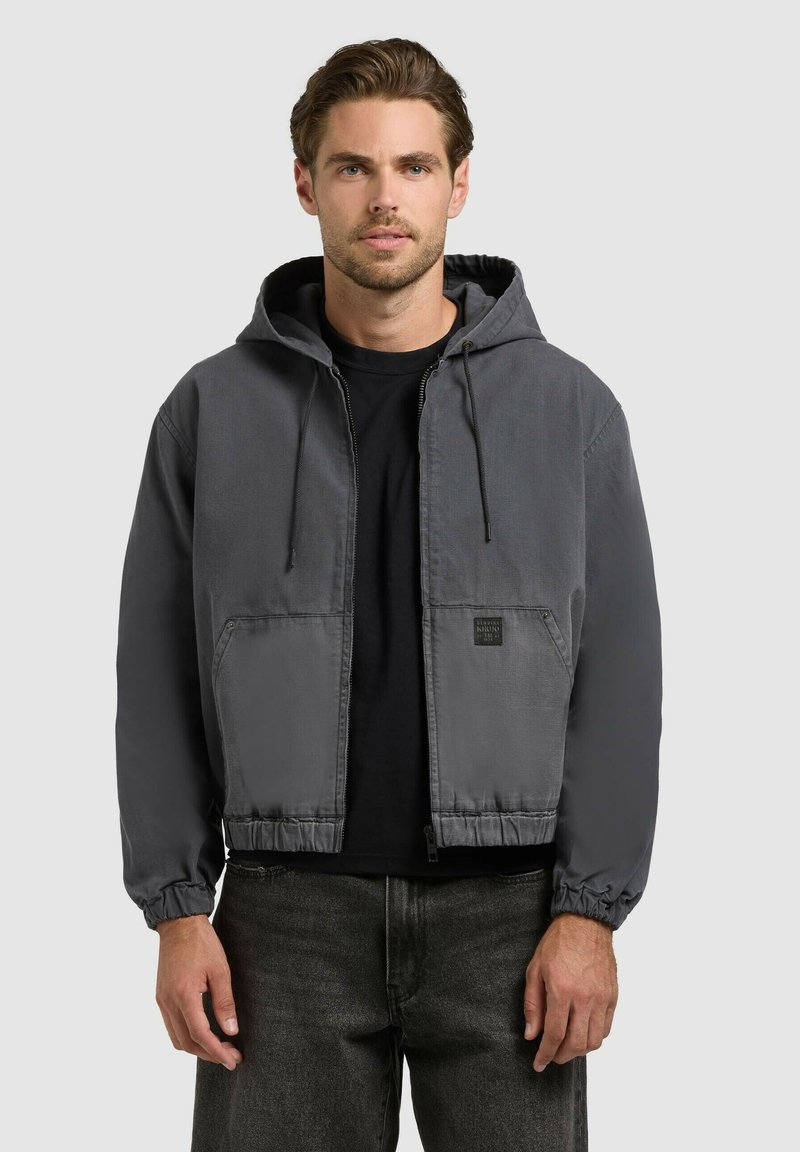 Homme portant une veste à capuche gris charbon avec des poches avant, sur un t-shirt noir et un jean foncé, debout devant un fond gris clair.