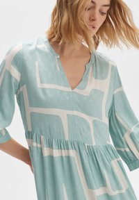 Hellblaues Kleid mit weißem geometrischen Design, verfügt über einen V-Ausschnitt, dreiviertellange Ärmel und eine geraffte Taille für zusätzliches Volumen.