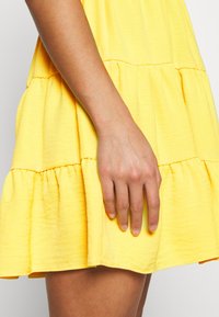 Žluté vrstvené šaty s texturovanou tkaninou, s nabraným lemem a přiléhavým živůtkem. Ruka je umístěna na sukni.