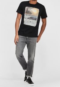 Camiseta de algodón negra con un gráfico de olas y un surfista, con el texto "O'Neill Surf". Llevada con jeans grises y zapatillas negras.