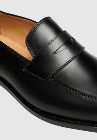 Mocassins en cuir noir avec un détail de sangle, texture lisse, accents cousus et bout arrondi, présentant une doublure intérieure contrastante.