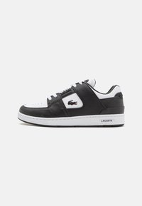 Baskets en cuir noir avec accents blancs, présentant une tige texturée, un embout perforé et le logo Lacoste sur le côté. Design sportif classique.