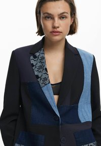 Tailored blazer featuring a mix of fabrics: dark navy, denim patches, and floral accents. Notable patchwork design and two buttons.