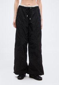 Άτομο που φοράει φαρδιά μαύρα νάιλον παντελόνια με κορδόνι στη μέση και μαύρα μπαλαρίνα, στέκεται μπροστά σε ένα απλό ανοιχτόφωτο φόντο.