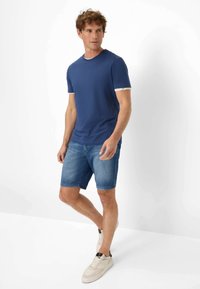 T-shirt blu a maniche corte con bordo bianco, abbinato a pantaloni corti in denim. Il modello indossa sneakers chiare. Design semplice, vestibilità casual e stile confortevole.