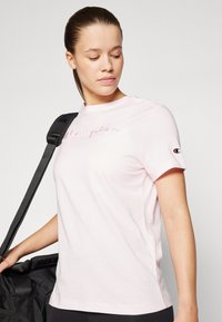 Lys rosa bomull t-skjorte med brodert "Champion" logo, rund hals, korte ermer, og et logo-lapp på ermet. Enkelt design.