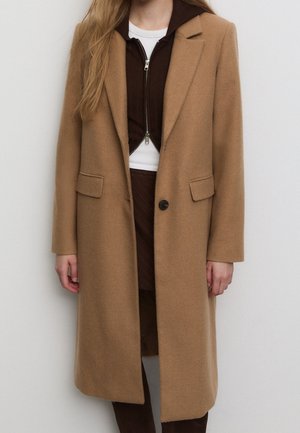 Femme portant un long manteau camel sur un haut marron foncé zippé et une jupe avec un sous-vêtement blanc, debout devant un fond uni.