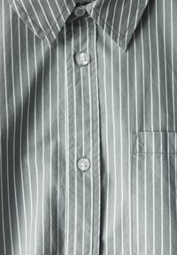 Vaaleanharmaa nappipaitapaita, jossa on pystysuoria valkoisia raitoja. Paidassa on klassinen kaulus, yksi rintatasku ja kirkkaat muoviset napit.