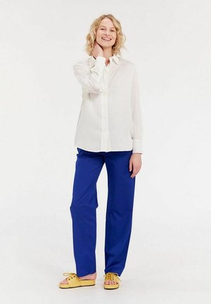 Chemise blanche à boutons, pantalon bleu et chaussures à enfiler jaunes. La tenue présente une coupe décontractée et une texture de tissu lisse.