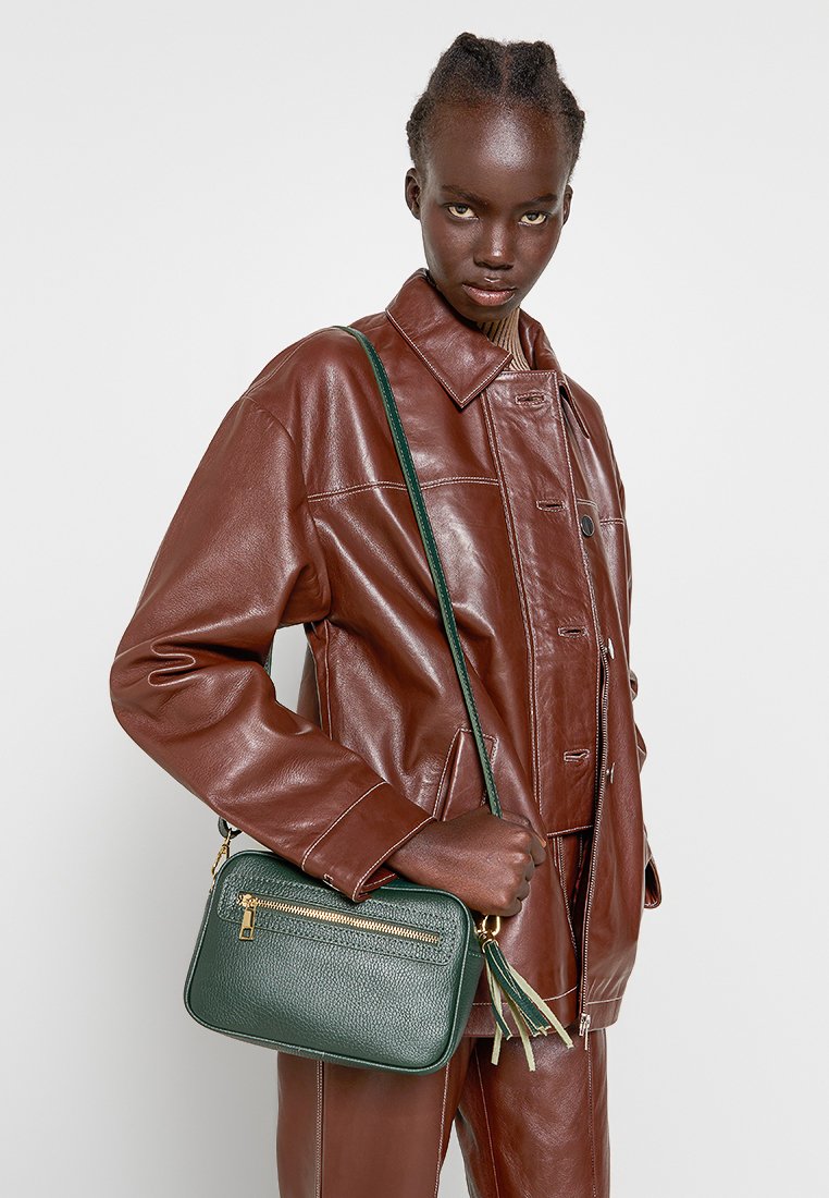 Sac en bandoulière en cuir vert avec une surface texturée, des finitions en or, et un compartiment zippé, tenu par un modèle portant une veste en cuir marron.