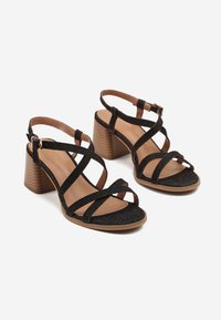 Sandales à talons noires avec des lanières croisées, dotées d'un dessus en tissu texturé et d'un talon bloc marron. Bride de cheville ajustable pour un maintien sûr.