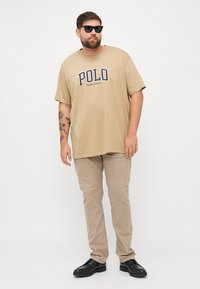 Bež oversize majica s napisom "POLO", u kombinaciji s svijetlosmešavim hlačama i crnim cipelama. Model nosi tamne sunčane naočale.