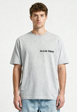 Jeune homme aux cheveux bruns bouclés porte un t-shirt gris clair avec le texte noir "YOU & ME TONIGHT" sur la poitrine gauche, assorti à un jean bleu clair.