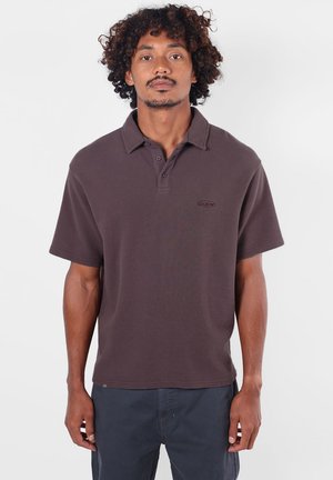 Homme aux cheveux bouclés portant un polo à manches courtes marron foncé et un pantalon foncé, debout devant un fond clair uni.