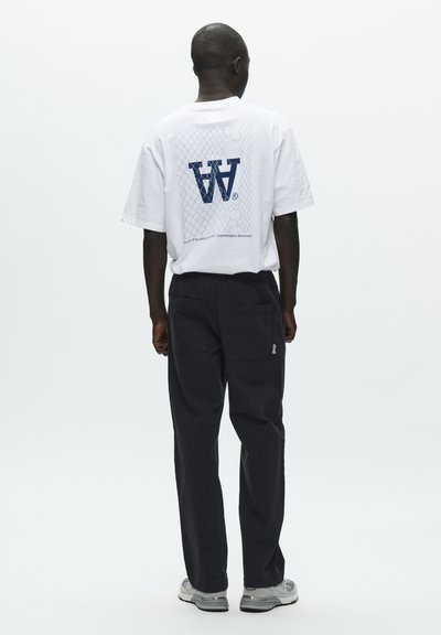T-shirt blanc à manches courtes avec un motif graphique bleu et un motif en résille au dos, associée à un pantalon ample foncé et des baskets grises.