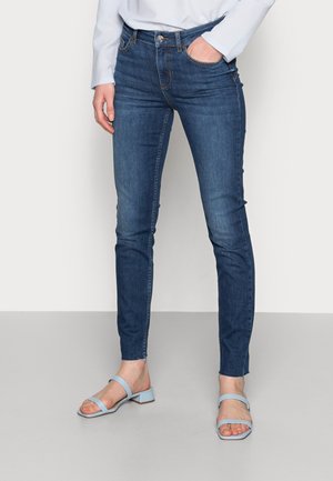Nainen, joka käyttää tummansinisiä skinny-farkkuja raaoilla helmillä ja vaaleansiniset avonaiset sandaalit matalilla lohkokoroilla.