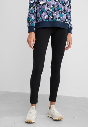 Person wearing black leggings, a floral patterned sweatshirt in purple and teal, and white sneakers with beige soles, standing on a gray floor.