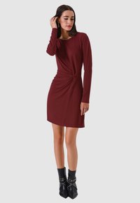 Abito bordeaux a maniche lunghe con dettaglio twist in vita, realizzato in materiale morbido; abbinato a stivaletti neri con dettagli borchiati.