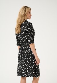 Musta mekko, jossa on valkoisia pilkkuja, lyhyet hihat, vyöllä varustettu vyötärö ja polveen ulottuva helma. Silkkinen kangas ja rento malli.
